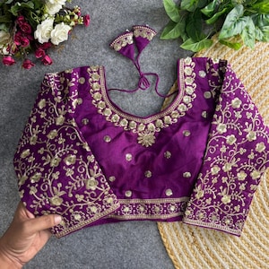 Puede incluir: Una blusa morada con bordados dorados. La blusa presenta mangas largas y un escote redondo, ambos adornados con intrincados diseños florales. El corpiño tiene un diseño floral central y pequeños detalles dorados. La blusa tiene un par de borlas decorativas a juego.