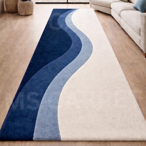 May include: A long, rectangular rug with a wavy design in shades of blue and white. The rug features a dark blue border that transitions into lighter shades of blue, culminating in a white central section. The rug is placed on a wooden floor.