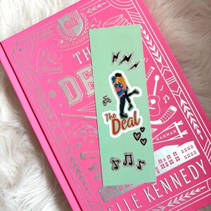 Könnte beinhalten: Ein pinkfarbenes Hardcover-Buch mit dem Titel "The Deal" von Elle Kennedy, mit einem hellgrünen Lesezeichen mit einem sich umarmenden Paar und dem Buchtitel. Das Lesezeichen hat auch Noten, Herzen und Blitzgrafiken.