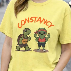 Op de afbeelding: Geel T-shirt met het woord "Constancy" in oranje letters. Het shirt heeft een cartoon schildpad en cactus karakter. De persoon draagt een blauwe spijkerbroek.
