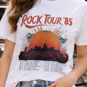 Puede incluir: Camiseta blanca con un gráfico de una puesta de sol y el texto "ROCK TOUR '85". La camiseta también enumera las fechas de la gira. La persona lleva pantalones cortos vaqueros azul claro.