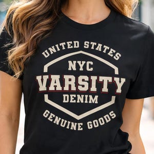 May include: Black t-shirt with a white and red graphic that reads "UNITED STATES NYC VARSITY DENIM GENUINE GOODS." The model is wearing blue jeans. The shirt is a crew neck and short-sleeved.
