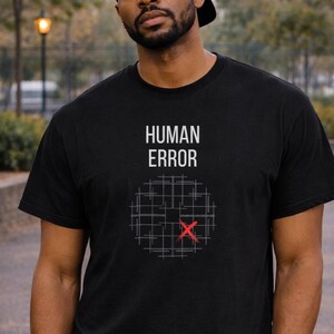 Op de afbeelding: Zwart T-shirt met de woorden "HUMAN ERROR" boven een rasterontwerp met een rode "X". De persoon op de afbeelding draagt een zwarte pet en een blauwe spijkerbroek.