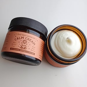 Peut inclure: Deux pots en verre brun de Calm Cream de Peace Roots Apothecary. L'un est ouvert, révélant un tourbillon de crème blanche. L'autre est fermé avec un couvercle noir. L'étiquette indique "Calm Cream" et "For Hormonal Harmony & A Calm Body."