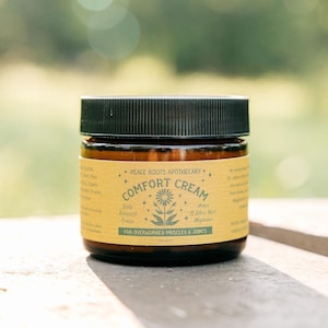 Puede incluir: Un frasco de vidrio marrón con tapa negra, etiquetado "Comfort Cream" con texto que indica que es para músculos y articulaciones sobrecargados. La etiqueta presenta un diseño floral y las palabras "Peace Roots Apothecary."
