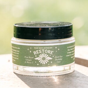Puede incluir: Un tarro de vidrio transparente con tapa negra, etiquetado "RESTORE" con un diseño de margarita. La etiqueta también dice "Plant Based Collagen & Vitamin C" y "Infused Face Decolletage Cream". El tarro es de 50 ml.