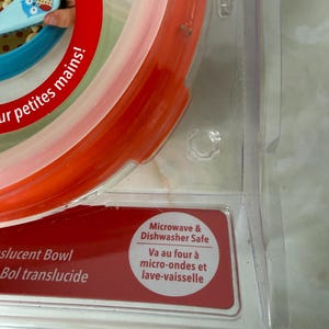 May include: A translucent bowl with red, white, and blue sections. The packaging includes the text "Microwave & Dishwasher Safe". The bowl is designed for small hands.