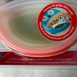 May include: A baby feeding set with a divided plate, removable training rim, and translucent bowl. The set includes a blue bowl with an owl design and a matching spoon. The packaging features the text "Training rim guides little hands!"