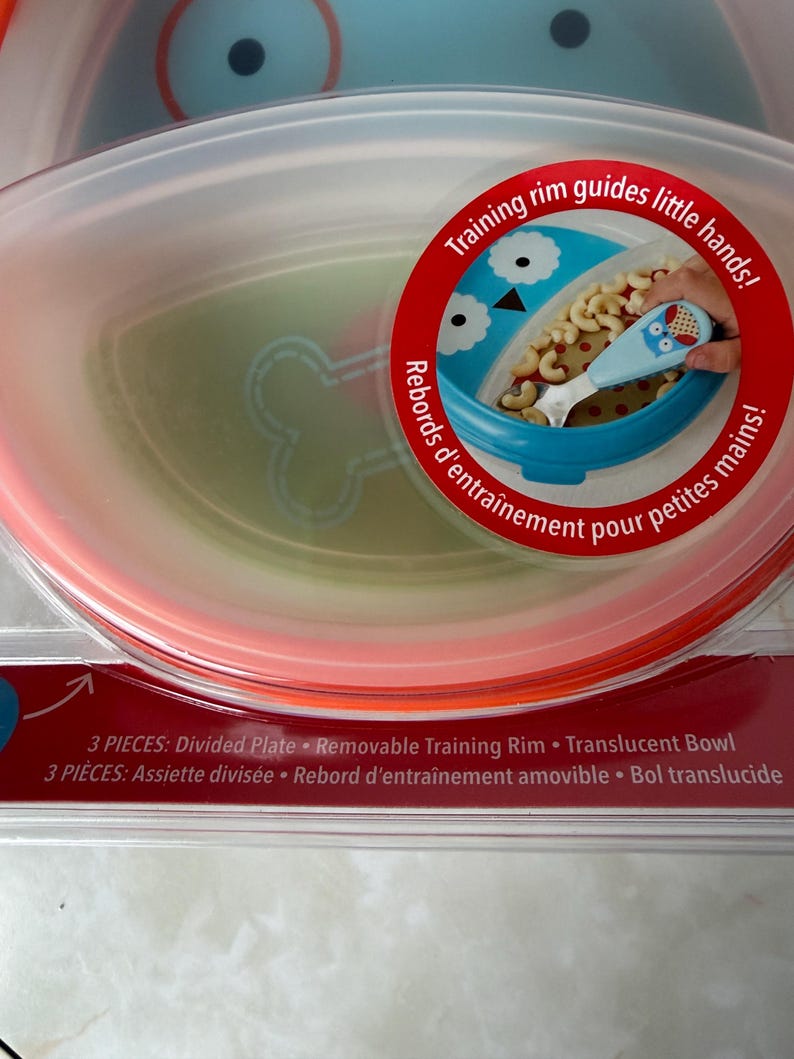 May include: A baby feeding set with a divided plate, removable training rim, and translucent bowl. The set includes a blue bowl with an owl design and a matching spoon. The packaging features the text "Training rim guides little hands!"