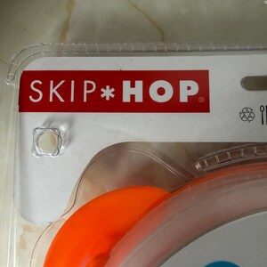 May include: Close-up of a baby spoon in its original packaging. The packaging features the brand name "SKIP*HOP" in white letters on a red background. The spoon is orange and designed for babies aged 6 months and older.