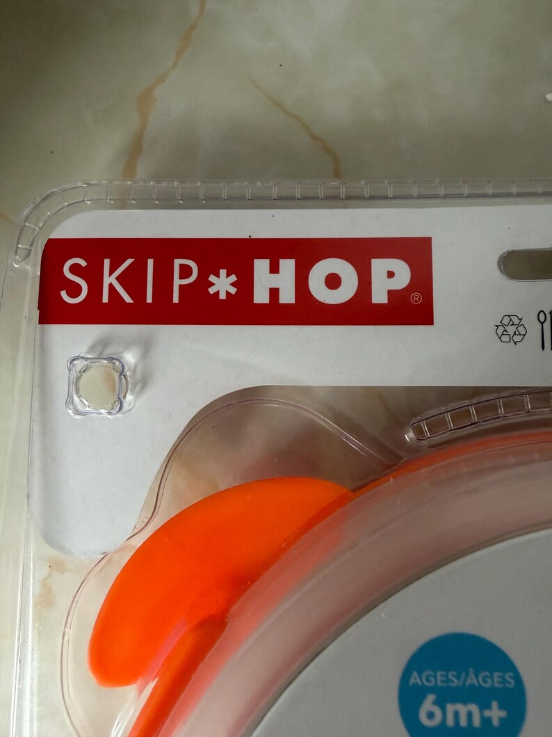May include: Close-up of a baby spoon in its original packaging. The packaging features the brand name "SKIP*HOP" in white letters on a red background. The spoon is orange and designed for babies aged 6 months and older.