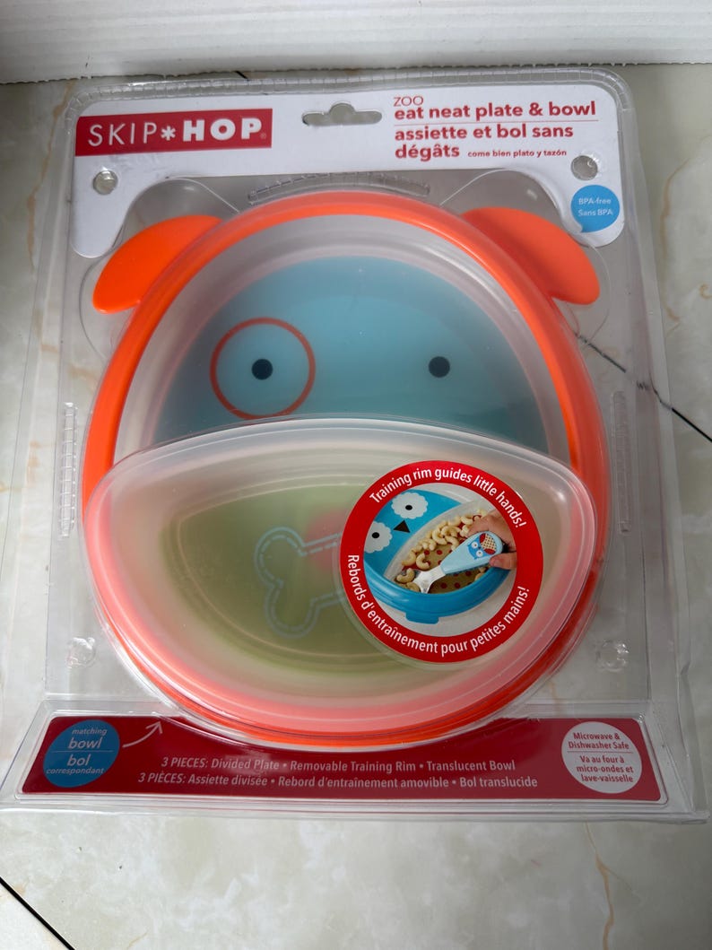 May include: A Skip Hop ZOO eat neat plate & bowl set in a clear plastic package. The set includes a divided plate, removable training rim, and translucent bowl. The bowl is blue with orange accents and a dog face design.