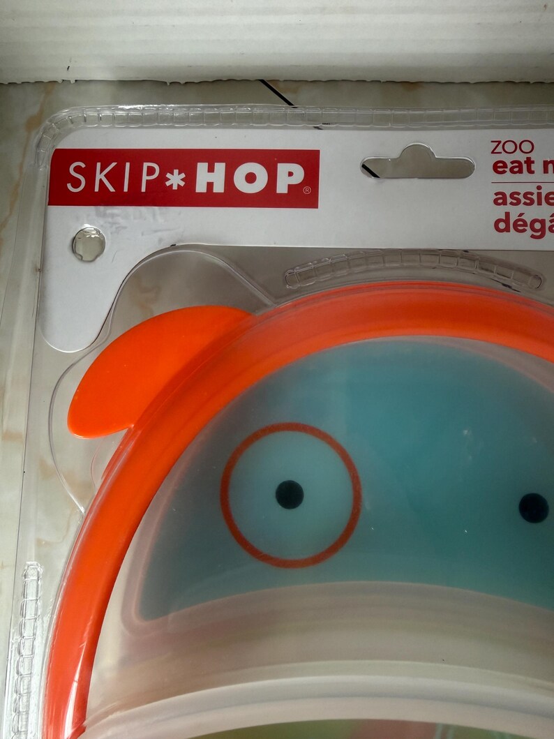 May include: A Skip Hop ZOO baby bowl with an orange rim and a blue interior. The bowl has a cartoon-like design with a large eye and a small orange fin. The packaging includes the Skip Hop logo in red.