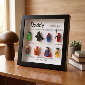 Op de afbeelding: Een zwarte schaduwdooslijst met acht superhelden-minifiguren in twee rijen. De tekst luidt: Daddy YOU ARE... STRONGER THAN, COOLER THAN, QUICKER THAN, FUNNIER THAN, SMARTER THAN, FASTER THAN, BRAVER THAN, TOUGHER THAN. Love, Mila & Falon.