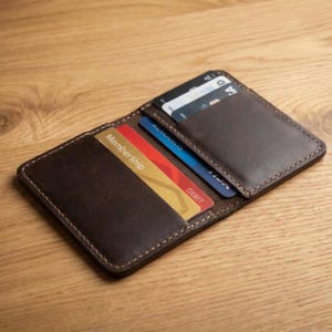 Peut inclure: Portefeuille en cuir marron ouvert, avec des coutures visibles, contenant plusieurs cartes de crédit et de membre. Le portefeuille est présenté ouvert, mettant en valeur ses poches intérieures et les cartes qu'il contient. Fond en bois.