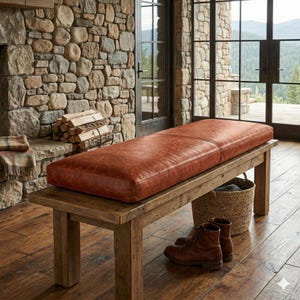 May include: A rustic wooden bench with a rectangular, reddish-brown leather cushion. The bench is placed on a wooden floor, with a woven basket and brown leather boots nearby. The background features a stone wall and large windows.