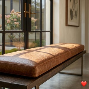 May include: A long, rectangular bench with a brown leather cushion and a dark wooden base. The bench is approximately 152 cm long and is placed near a large window, with sunlight illuminating the leather.