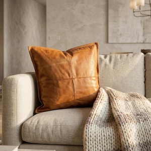 May include: A square, tan leather pillow with stitched detailing rests on a light-colored sofa. A chunky knit blanket in shades of cream and brown is draped over the sofa. The pillow has a warm, inviting appearance, perfect for home decor.