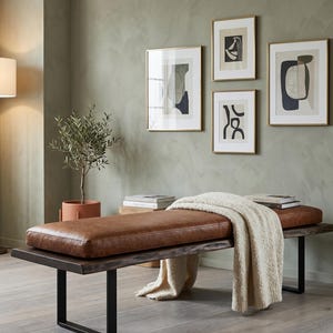 May include: A rectangular bench with a brown leather-like cushion and a dark wood base featuring black metal legs. A cream-colored textured throw blanket is draped over the seat. The bench is positioned in a room with four framed abstract art prints.