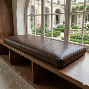 May include: A rectangular dark brown leather bench cushion with smooth, taut upholstery and clean, straight edges. The cushion is positioned horizontally on a wooden surface, featuring a consistent, matte finish across its entire top surface.