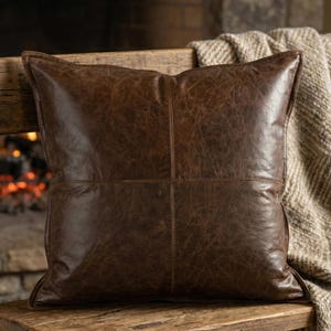 May include: A square, brown leather pillow with a textured surface and stitched details. The pillow is placed on a wooden bench with a neutral-colored blanket draped over it. The background features a fireplace.