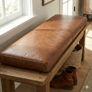 Può includere: Una panca in pelle marrone con un cuscino rettangolare poggia su una struttura in legno rustico. La panca è vicino a una finestra, con un cesto intrecciato e stivali in pelle marrone sotto. La panca è lunga circa 152 cm.