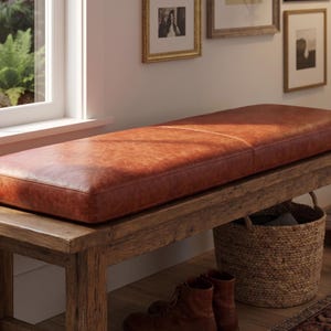 May include: A rectangular bench featuring a thick, smooth, cognac-colored leather cushion resting on a sturdy, rustic wooden frame with four legs. The leather surface displays natural grain variations and a warm, polished finish.