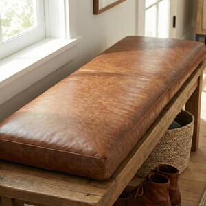 May include: A rectangular bench featuring a smooth, cognac-colored faux leather upholstered seat cushion. The bench is supported by a sturdy wooden frame with visible grain. A woven basket and a pair of brown leather boots sit underneath.
