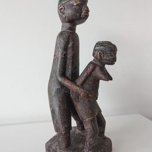 Könnte beinhalten: Eine geschnitzte Holzskulptur, die zwei Figuren darstellt. Die größere Figur steht aufrecht, während die kleinere Figur davor positioniert ist. Die Skulptur ist überwiegend dunkelbraun mit sichtbarer Holzmaserung und Altersspuren.