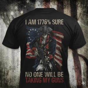 Könnte beinhalten: Ein schwarzes T-Shirt mit einer Grafik eines Soldaten in Kolonialuniform, der ein modernes Gewehr vor einer historischen amerikanischen Flagge hält. Der Text lautet I AM 1776% SURE NO ONE WILL BE TAKING MY GUNS in einer Vintage-Schrift.