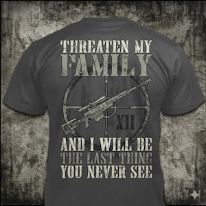 Può includere: T-shirt grigio scuro con la scritta "Threaten My Family And I Will Be The Last Thing You Never See." Un fucile con mirino è centrato su una grafica a bersaglio.