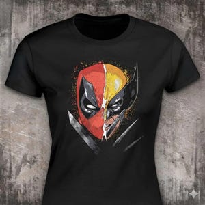 May include: A black short-sleeved t-shirt featuring a graphic print of a split mask design. The left side is red with black eye detailing, and the right side is yellow with black eye detailing. Sharp metallic blades are visible at the bottom.
