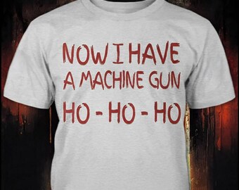 May include: A light gray short-sleeved t-shirt featuring red text printed on the front. The text reads, "NOW I HAVE A MACHINE GUN HO-HO-HO" in a distressed, hand-written style font. The shirt has a crew neckline and a standard fit.