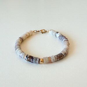 Peut inclure: Un bracelet composé de petites perles plates en forme de disque dans des tons variés de beige, crème, gris et marron. Le bracelet comporte une perle centrale en métal doré brillant et se ferme à l'aide d'un fermoir mousqueton de couleur dorée.