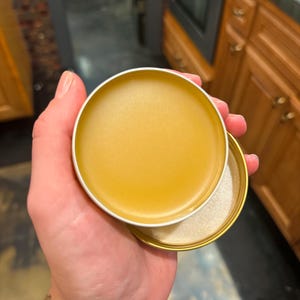 May include: A small, open, round, gold-colored tin containing a smooth, yellow balm. The lid is off, revealing the balm and a white, shimmering substance. The tin is held in a hand.