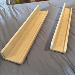 May include: Two unfinished wooden shelves with a U-shaped design. The shelves are made of light-colored wood and are rectangular with a raised edge on one side. They are placed on a gray fabric surface.