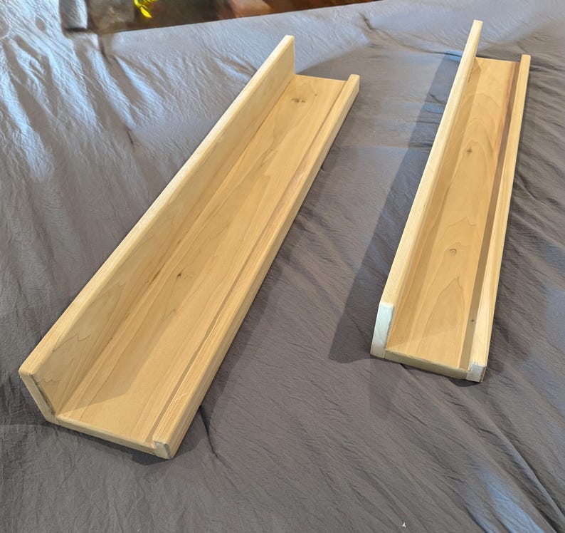 May include: Two unfinished wooden shelves with a U-shaped design. The shelves are made of light-colored wood and are rectangular with a raised edge on one side. They are placed on a gray fabric surface.