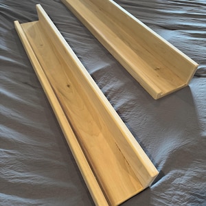 May include: Two unfinished wooden shelves with a trough-like design. The shelves are made of light-colored wood and have a rectangular shape with raised sides. They are placed on a gray fabric surface.
