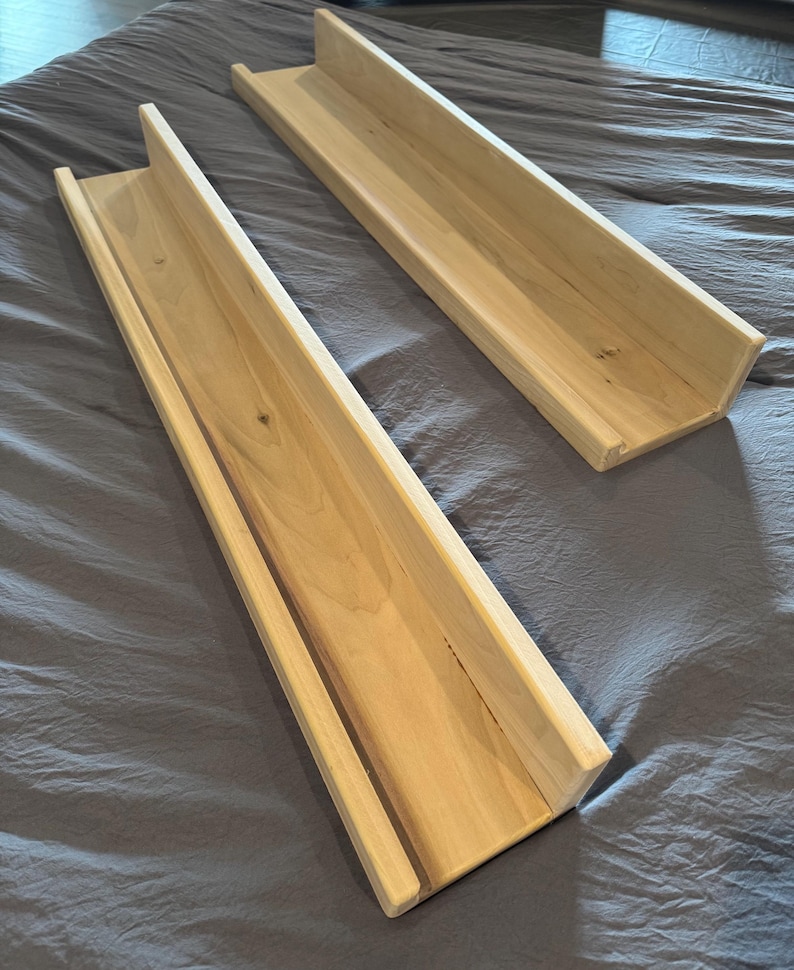 May include: Two unfinished wooden shelves with a trough-like design. The shelves are made of light-colored wood and have a rectangular shape with raised sides. They are placed on a gray fabric surface.
