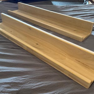 May include: Two unfinished wooden floating shelves. The shelves are rectangular with a raised edge on three sides. The wood has a light, natural color and visible grain. The shelves are approximately 36 inches long.