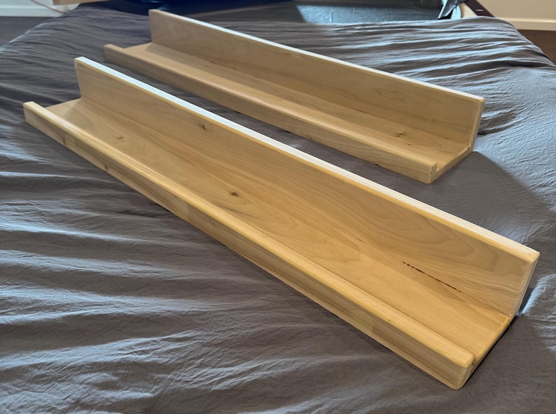 May include: Two unfinished wooden floating shelves. The shelves are rectangular with a raised edge on three sides. The wood has a light, natural color and visible grain. The shelves are approximately 36 inches long.