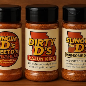May include: Three spice jars with black lids and labels featuring the text "SLINGIN' the D's SWEET D'S HONEY HEAT", "DIRTY D's CAJUN KICK", and "SLINGIN' the D's RUB-SOME D'SNT". The jars are filled with a reddish-brown spice blend.