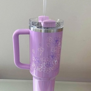 May include: A lavender insulated tumbler with a handle, clear lid, and straw. The tumbler features a floral design in white and purple. The handle and lid are the same lavender color as the body of the tumbler.