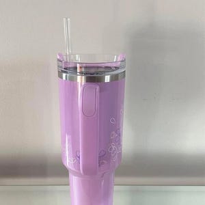 May include: A lavender insulated tumbler with a clear lid and a clear straw. The tumbler has a handle and is decorated with a floral design. The tumbler is designed to hold cold beverages.