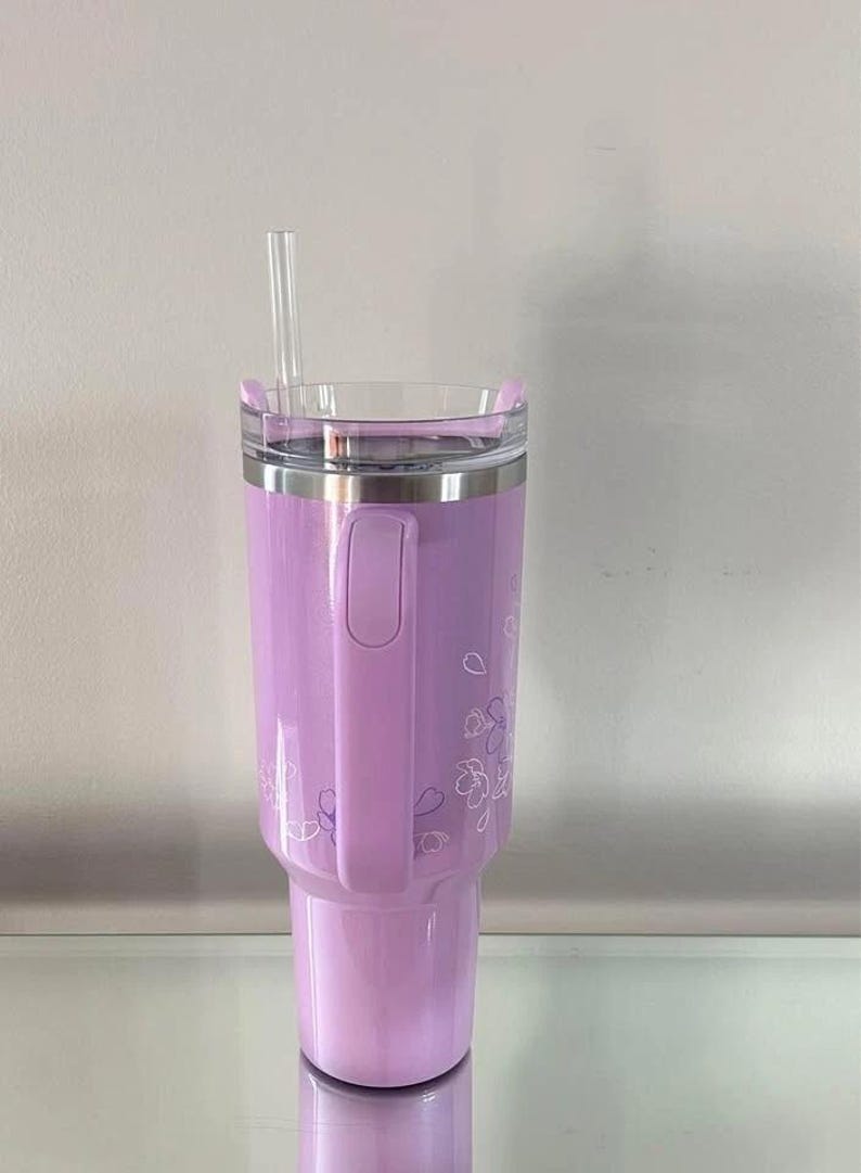 May include: A lavender insulated tumbler with a clear lid and a clear straw. The tumbler has a handle and is decorated with a floral design. The tumbler is designed to hold cold beverages.