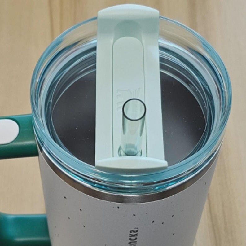 K&ouml;nnte beinhalten: Ein Edelstahl-Reisebecher mit hellgr&uuml;nem Deckel und Griff. Der Becher hat einen durchsichtigen Strohhalm und einen hellgr&uuml;nen Schieber zum Verschlie&szlig;en des Trinklochs. Der Becher hat ein gesprenkeltes Design.
