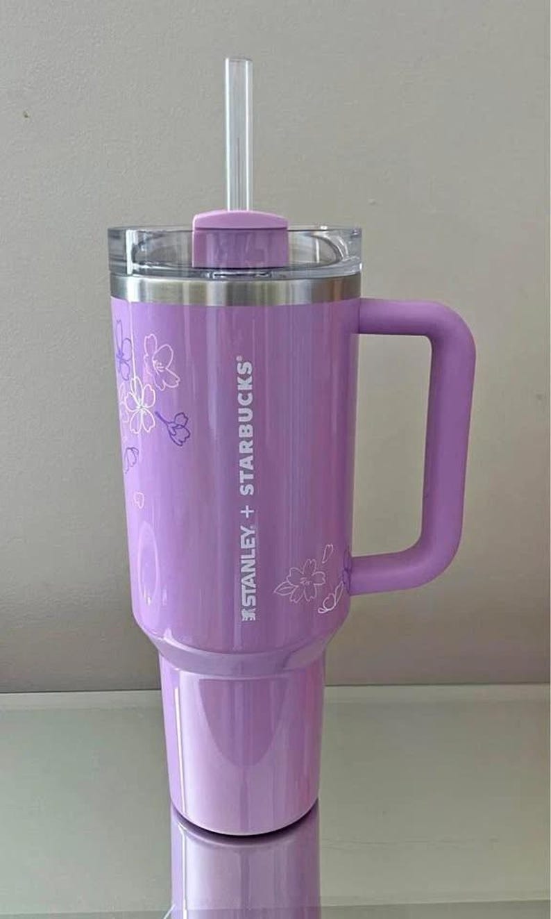 May include: A lavender-colored Stanley + Starbucks tumbler with a handle and a clear straw. The tumbler features a floral design and the text "STANLEY + STARBUCKS". The lid is clear, and the handle is the same lavender color as the body.