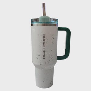 K&ouml;nnte beinhalten: Ein wei&szlig;er und gesprenkelter Stanley + Starbucks Becher mit gr&uuml;nem Griff und Deckel. Er hat einen silbernen Strohhalm und den Text "STANLEY + STARBUCKS" vertikal aufgedruckt. F&uuml;r kalte Getr&auml;nke konzipiert.