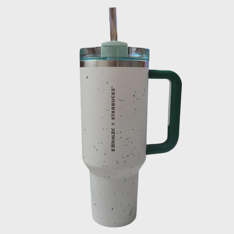 K&ouml;nnte beinhalten: Ein wei&szlig;er und gesprenkelter Stanley + Starbucks Becher mit gr&uuml;nem Griff und Deckel. Er hat einen silbernen Strohhalm und den Text "STANLEY + STARBUCKS" vertikal aufgedruckt. F&uuml;r kalte Getr&auml;nke konzipiert.