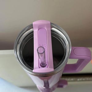 May include: A lavender-colored travel mug with a clear straw and a matching handle. The mug has a clear lid with a sliding closure for the straw. The mug has the words "STAINLESS STEEL" printed on the side.